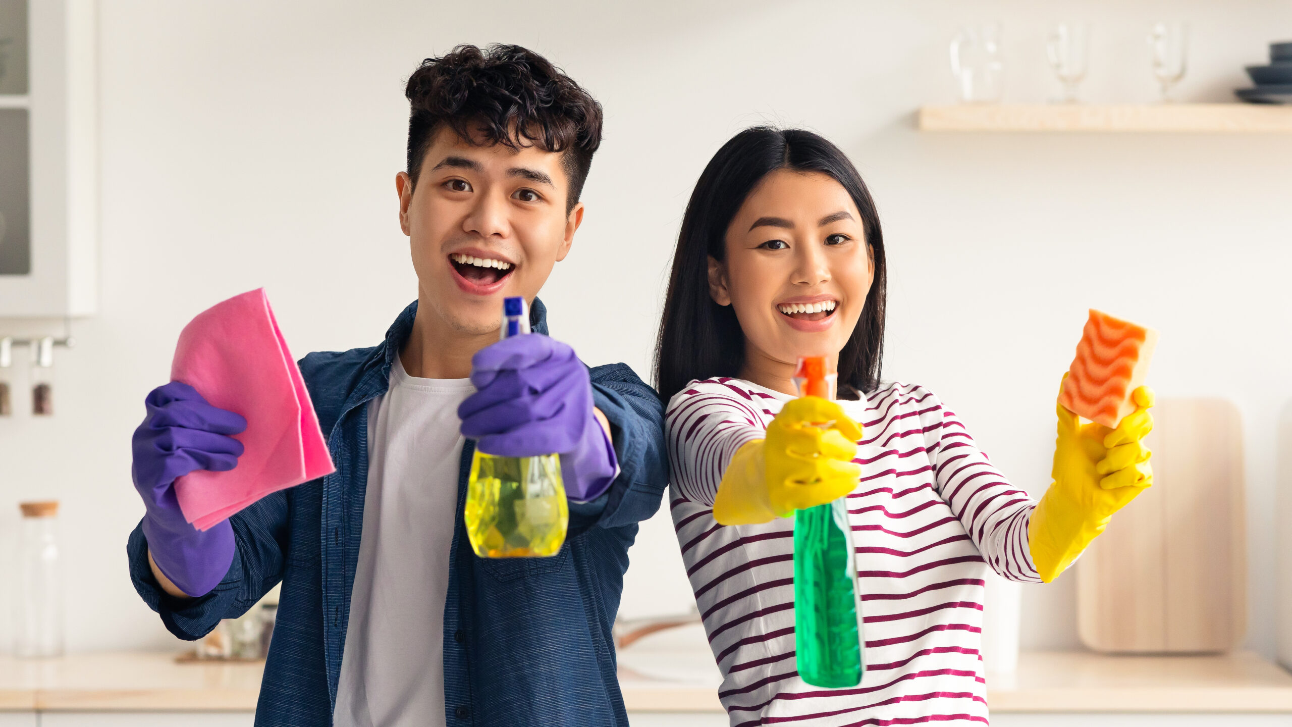 Cheerful asian couple with supplies for cleaning house posing in kitchen, funny young man and woman in colorful gloves standing together with spray bottles, house-keeping concept